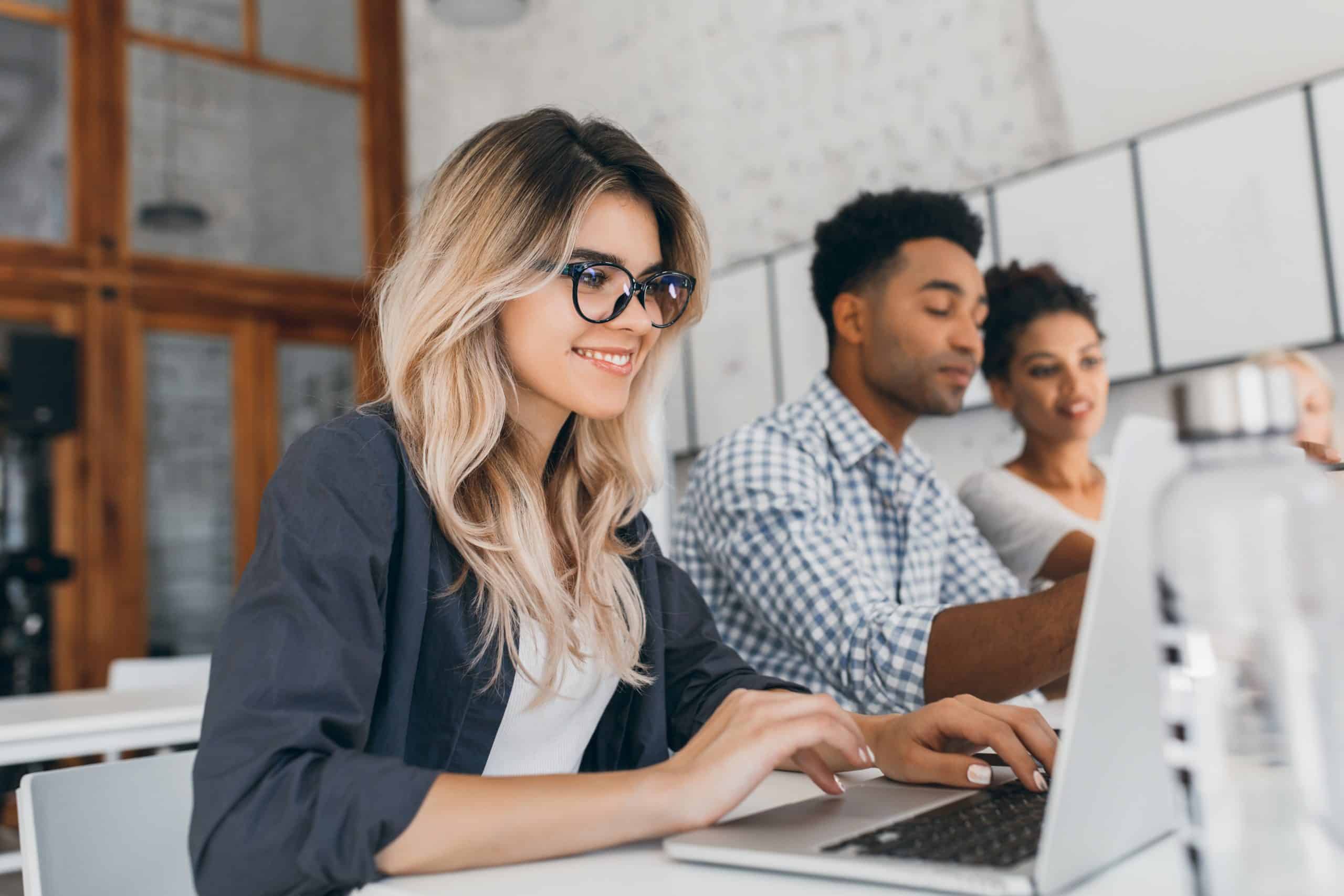 Beautiful curly female freelancer with cute manicure using laptop and smiling. Indoor portrait of blonde secretary sitting beside african coworker in blue shirt..